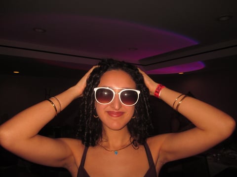 Woman wearing white sunglasses and gold bracelets posing with hands behind head in a nightclub setting with purple ceiling lights
