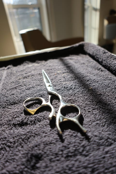 Ornate decorative scissors with silver handles resting on a gray textured towel, with window and doorway blurred in background