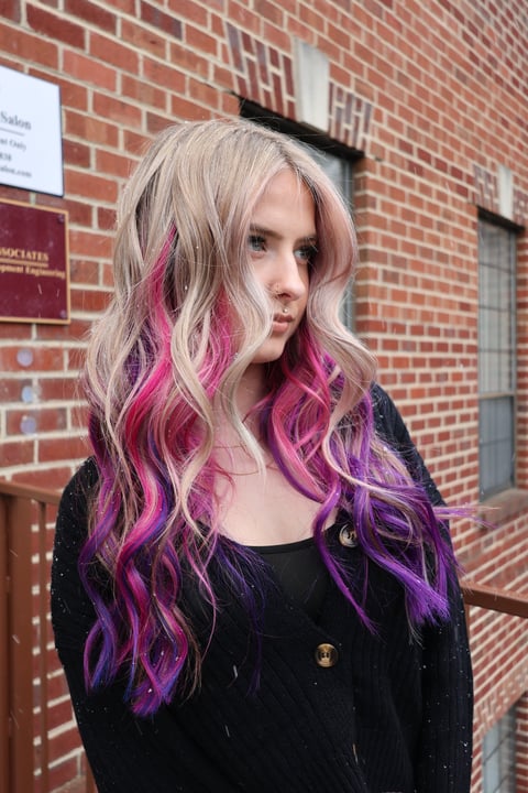 Young woman with long wavy blonde hair with vibrant pink and purple highlights wearing black top against red brick wall