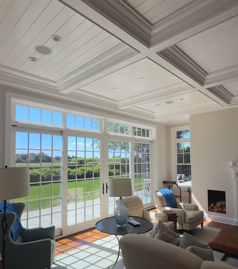 Bright modern living room with coffered ceiling, large glass doors and windows overlooking green lawn, neutral furnishings, and fireplace