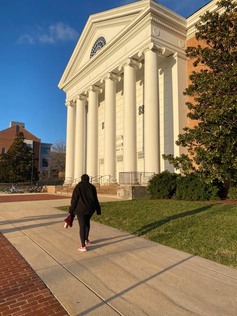 Person walking toward a classical white columned building with ivy-covered brick walls on a sunny day