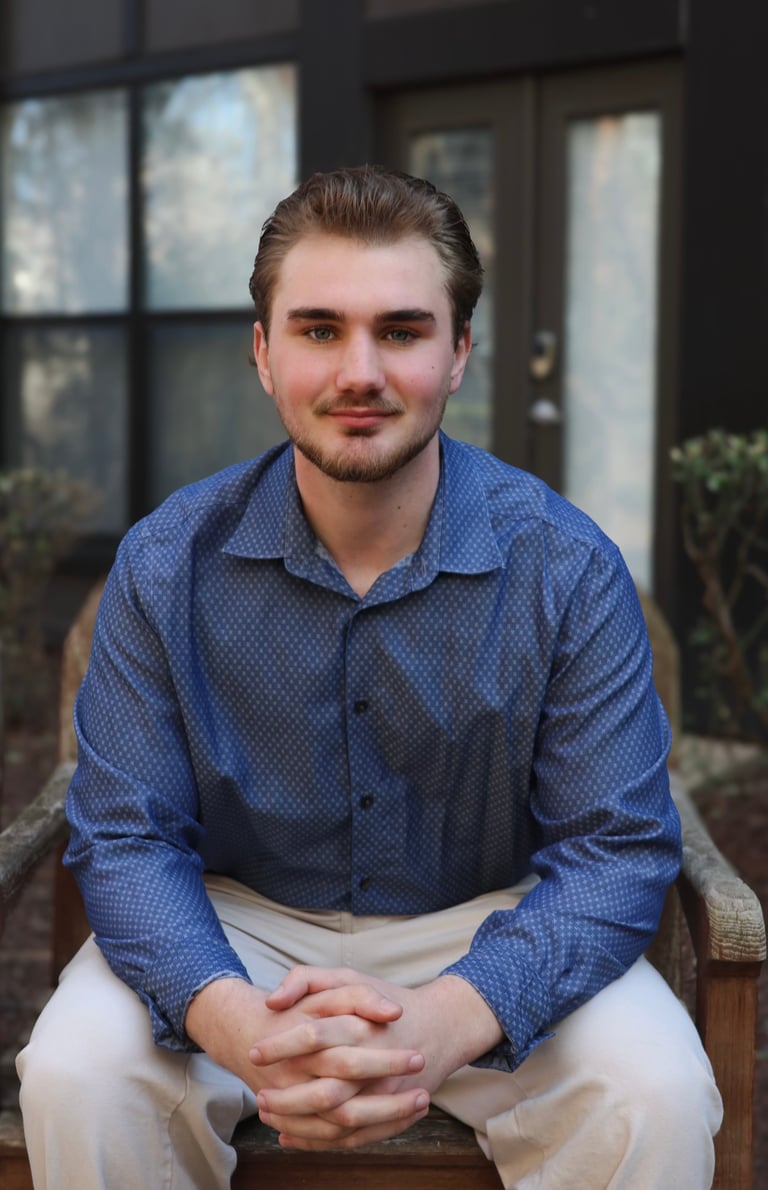 Young man sitting outdoors wearing a blue patterned button-up shirt and white pants, smiling at the camera